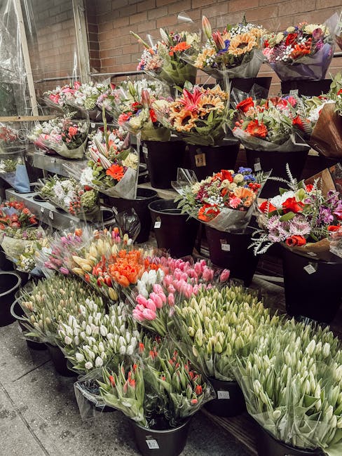 A display of various fresh flower bouquets arranged in black buckets and wrapping paper, including pink, white, red, orange, and purple blooms such as tulips, lilies, and chrysanthemums. The flowers a