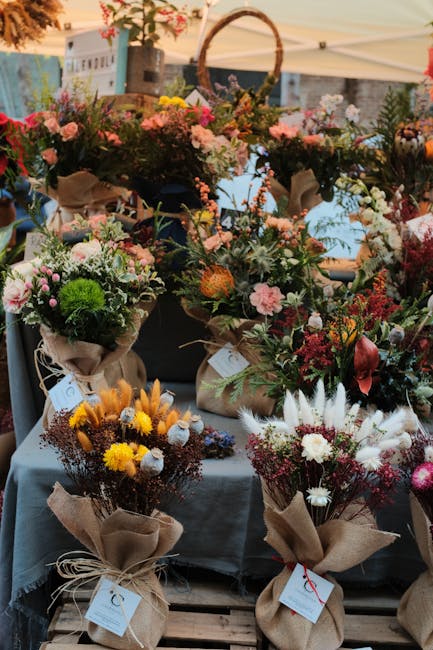 A vibrant display of floral arrangements at a flower market, featuring a variety of fresh flowers including pink and white roses, yellow mimosa, deep red blooms, and green foliage. The flowers are arr