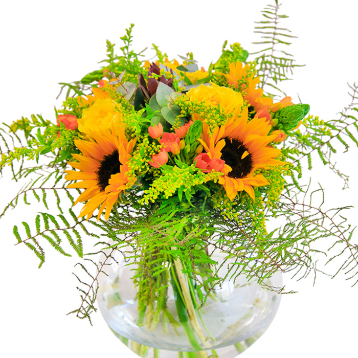 A first glance at this bouquet instantly recalls a stroll through Shoreditch's vibrant lanes at the peak of summer, every detail crafted with the precision and passion of a local expert. Sunflowers with their prominent brown centers and rich yellow petals stand tall as the focal point, sharing space with tightly-furled yellow garden roses whose soft, spiraled petals invite a closer look. Sprigs of red Hypericum berries are scattered among the blooms, each one a bright gem of colour that brings to mind the creative energy of Shoreditch's street art. The arrangement is lushly supported by textured layers of greenery: the cascading fronds of fern, light fillers like goldenrod or solidago with tiny yellow blossoms, and slender green stems that give structure and lightness. Eucalyptus leaves peek through in gentle blue-green, subtly cooling the palette much like the shady corners of Arnold Circus. Housed in an elegant, round glass vase, the stems crisscross below the flowers, visible in clear water, offering a glimpse into the skillful craftsmanship behind each element. The bouquet's scent is a soft mingling of rose petal and fresh grass, evoking memories of outdoor art fairs or summer gatherings. Designed for both visual and emotional impact, it's ideal for bringing Shoreditch's unique blend of energy and warmth into homes, perfect for birthdays, congratulations, or simply as a striking centerpiece that reflects the area's ever-present creativity.